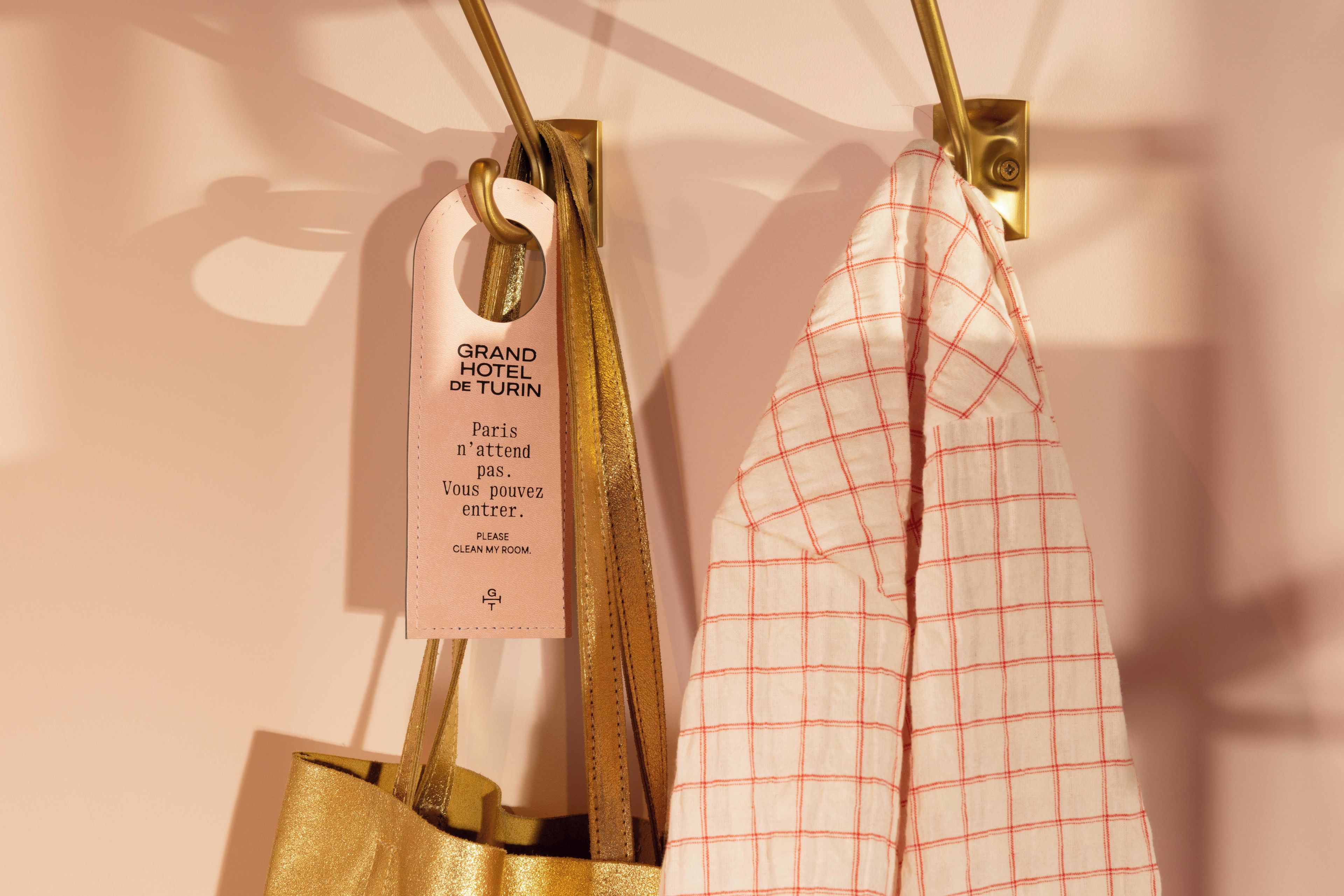 Twee tassen hangen aan een muur, waarvan er één een label heeft met de tekst 'GRAND HOTEL DE TURIN Parijs wacht niet. Kom binnen. MAAK MIJN KAMER schoon.'