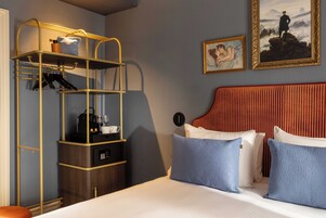 A hotel room with a bed, a bedside table with a lamp, a wall-mounted shelf, and two paintings.