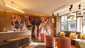 Un área de recepción moderna con un mostrador de madera, un mural, asientos con cojines naranja y vistas a la calle a través de grandes ventanales.