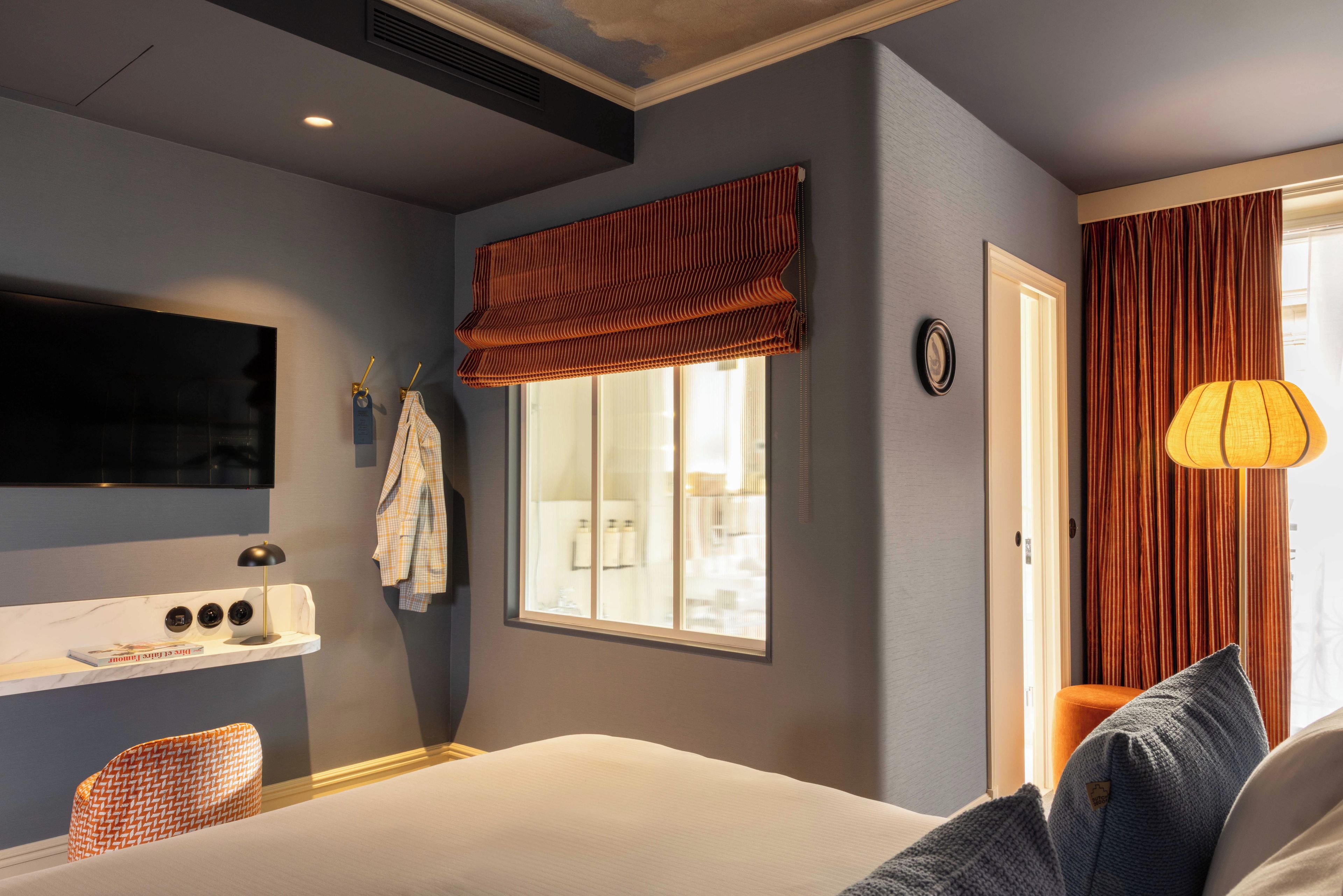 Habitación de hotel con cama, televisor montado en la pared, ventana con cortinas, mesita de noche con lámpara y vistas al exterior.