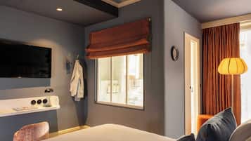 Habitación de hotel con cama, televisor montado en la pared, ventana con cortinas, mesita de noche con lámpara y vistas al exterior.