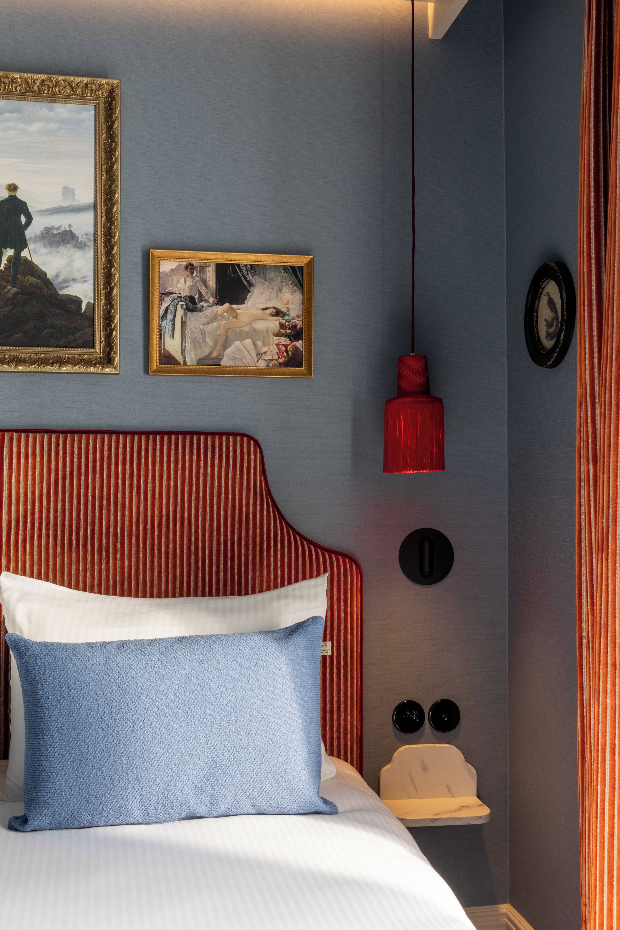 Een slaapkamer met een bed, twee ingelijste kunstwerken, een rode hanglamp en een klein nachtkastje met twee zwarte objecten.