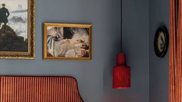 Een slaapkamer met een bed, twee ingelijste kunstwerken, een rode hanglamp en een klein nachtkastje met twee zwarte objecten.