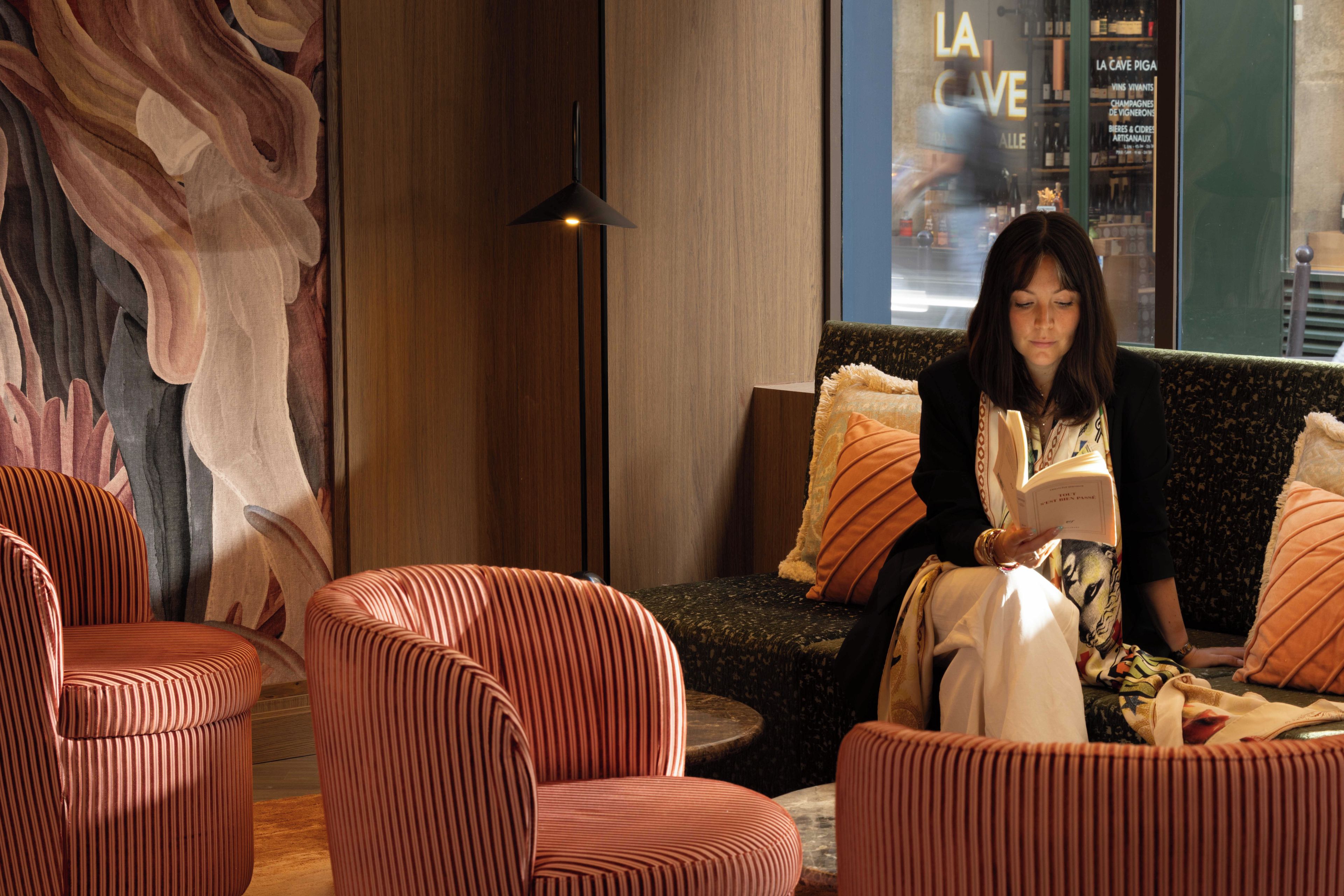 A person seated on a sofa in a room with striped chairs, a large abstract painting on the wall, and a window with a view of a store named 'LA CAVE'.