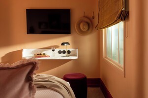 A bedroom with a bed, a bedside table with a lamp, a TV mounted on the wall, and a window with a curtain.