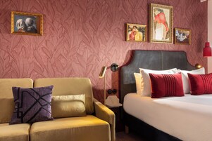 Una camera d'albergo con un divano, un letto e un comodino con una lampada. Alle pareti, si vedono quadri incorniciati.