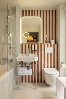 A bathroom with a glass-enclosed shower, a white sink with chrome fixtures, a toilet, and a striped wall with a mirror above the sink.