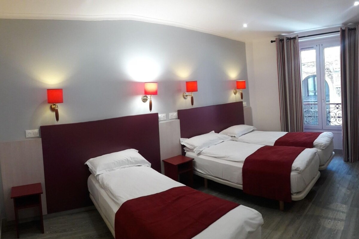 A hotel room with three beds, each with white bedding and red accents, and wall-mounted lamps.