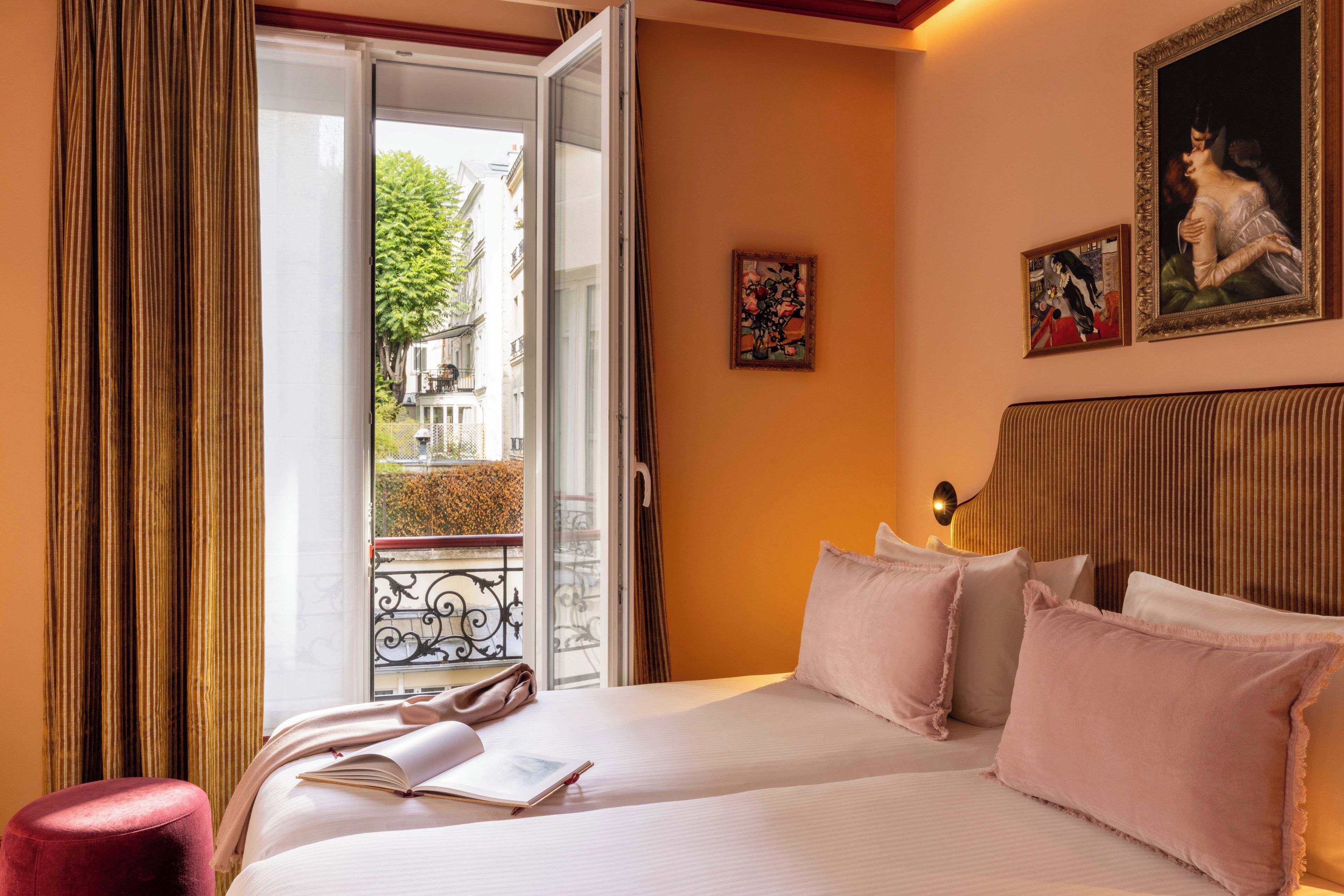 A hotel room with a bed, a window with curtains, a view of the street, and two paintings on the wall.