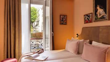 Une chambre d’hôtel avec un lit, une fenêtre avec des rideaux, une vue sur la rue et deux tableaux accrochés au mur.