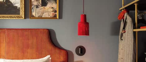 Une chambre d’hôtel avec un lit, un lampadaire rouge suspendu, un porte-manteau et deux tableaux au mur.