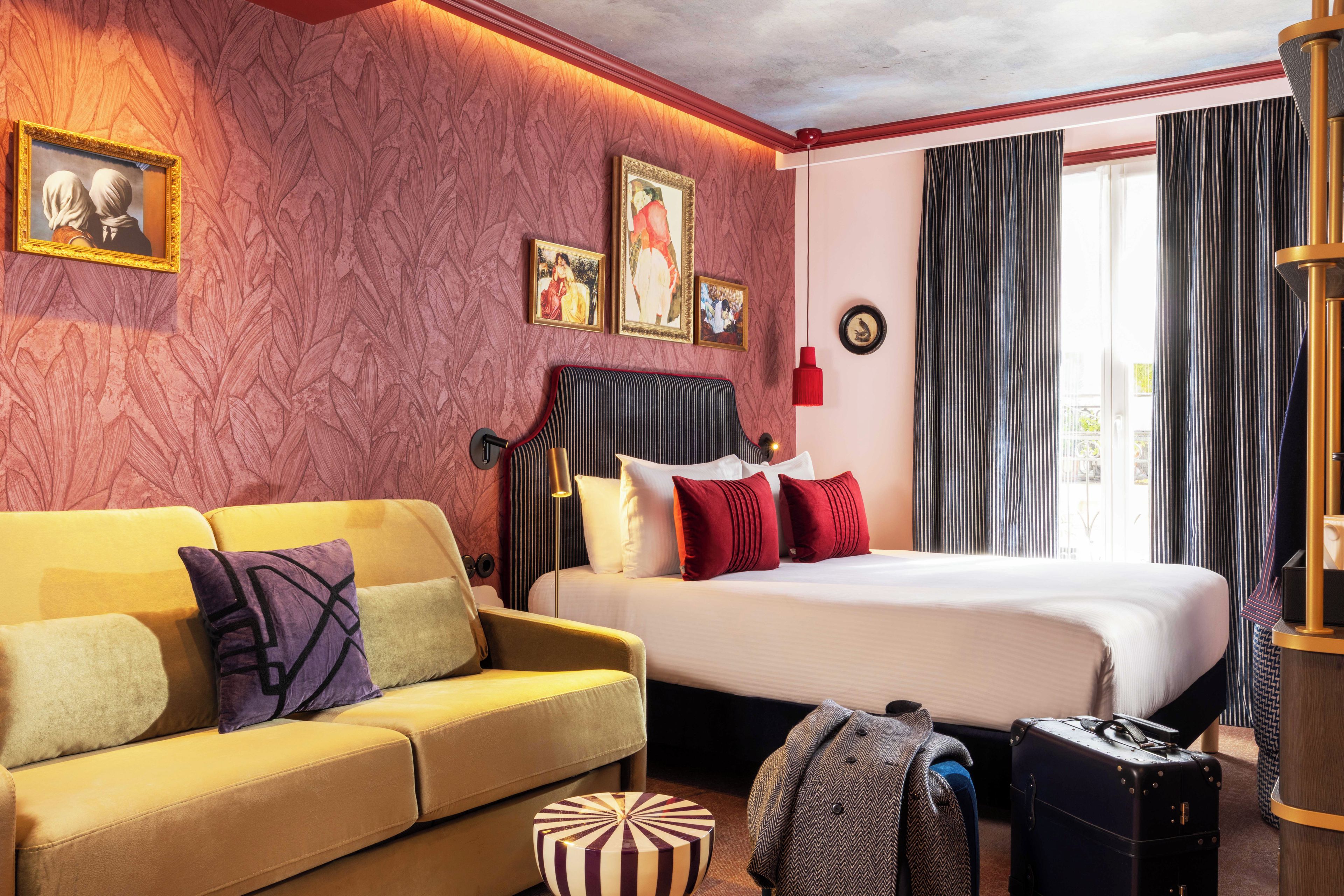 Une chambre d’hôtel avec un lit, un canapé, un petit pouf rond et une valise.