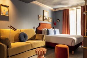 Una camera d'albergo con un divano, un letto, un poggiapiedi e una finestra con tende.
