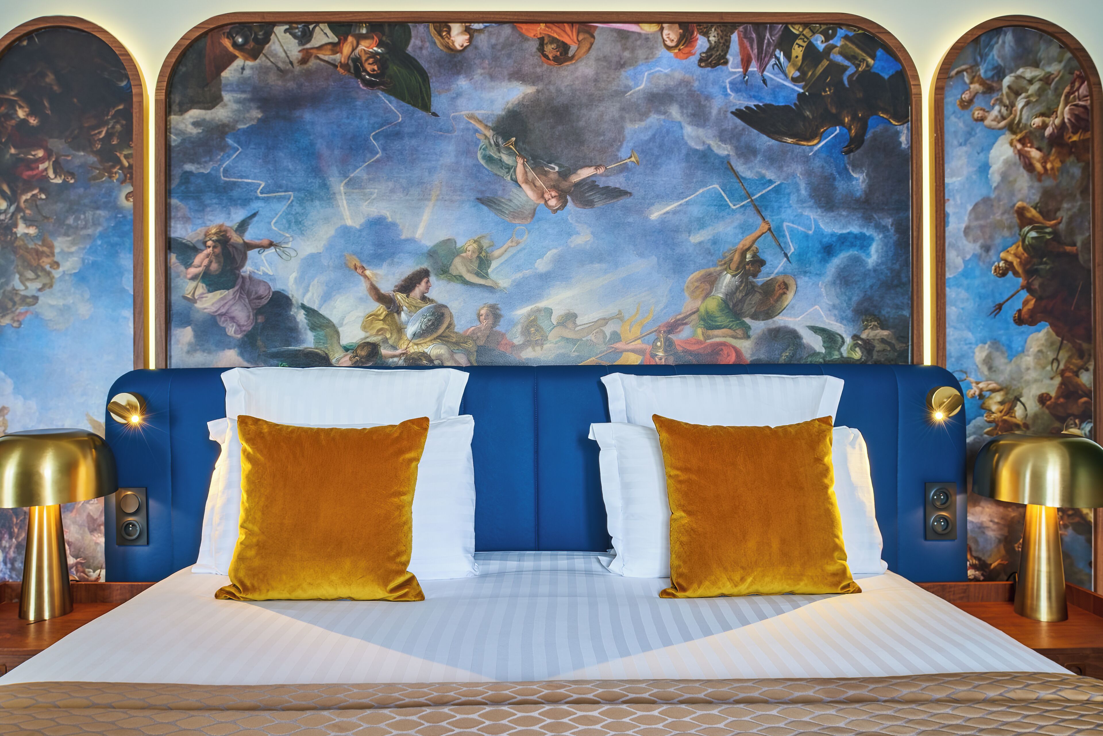 A bed with a blue headboard, white and gold pillows, and a mural of mythological figures on the wall behind.