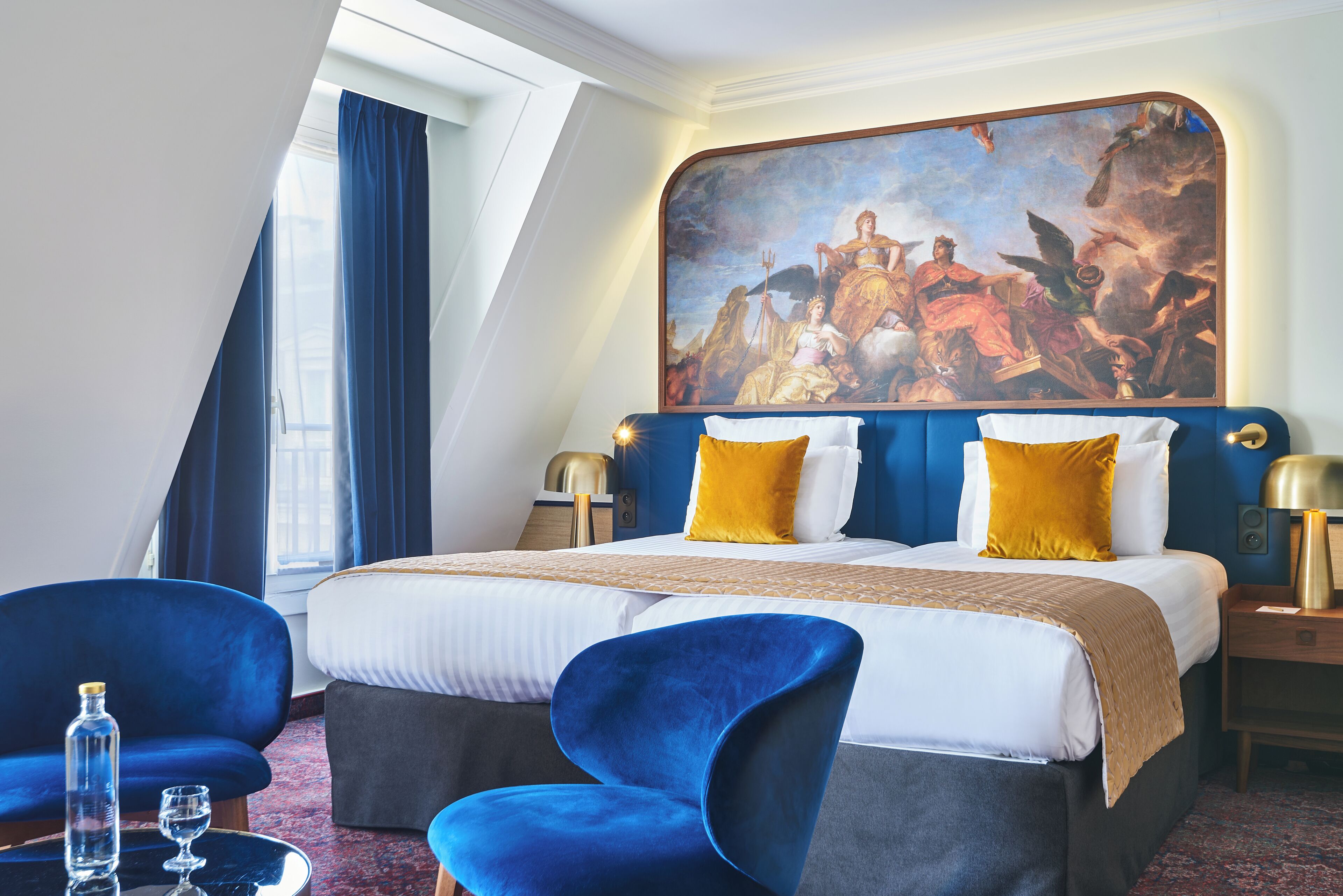 Habitación de hotel con una cama grande, dos sillones azules, un cuadro sobre la cama y una ventana con cortinas.