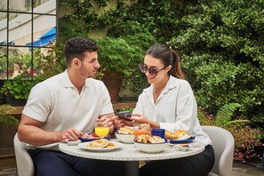 Two people seated at a round table outdoors, enjoying breakfast with plates of food, drinks, and a smartphone. - Hotel Le Versailles (Versailles)