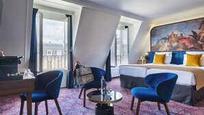 Une chambre d’hôtel avec un grand lit, deux fauteuils bleus, une table ronde avec une bouteille et un verre, et un tableau au-dessus du lit.