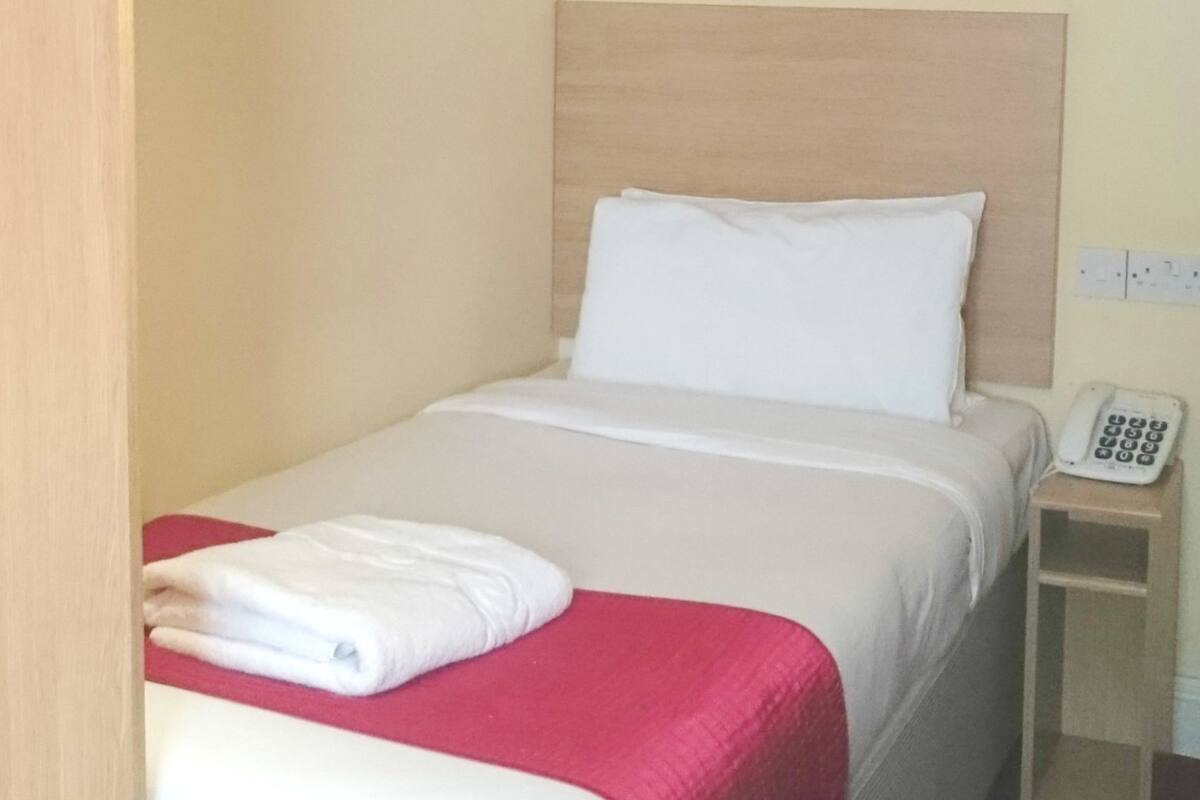 A single bed with a wooden headboard, a white pillow, and a red blanket. There is a small bedside table with a telephone.