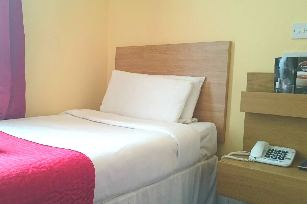 A hotel room with a bed, a bedside phone, and a shelf with a bottle and a glass.