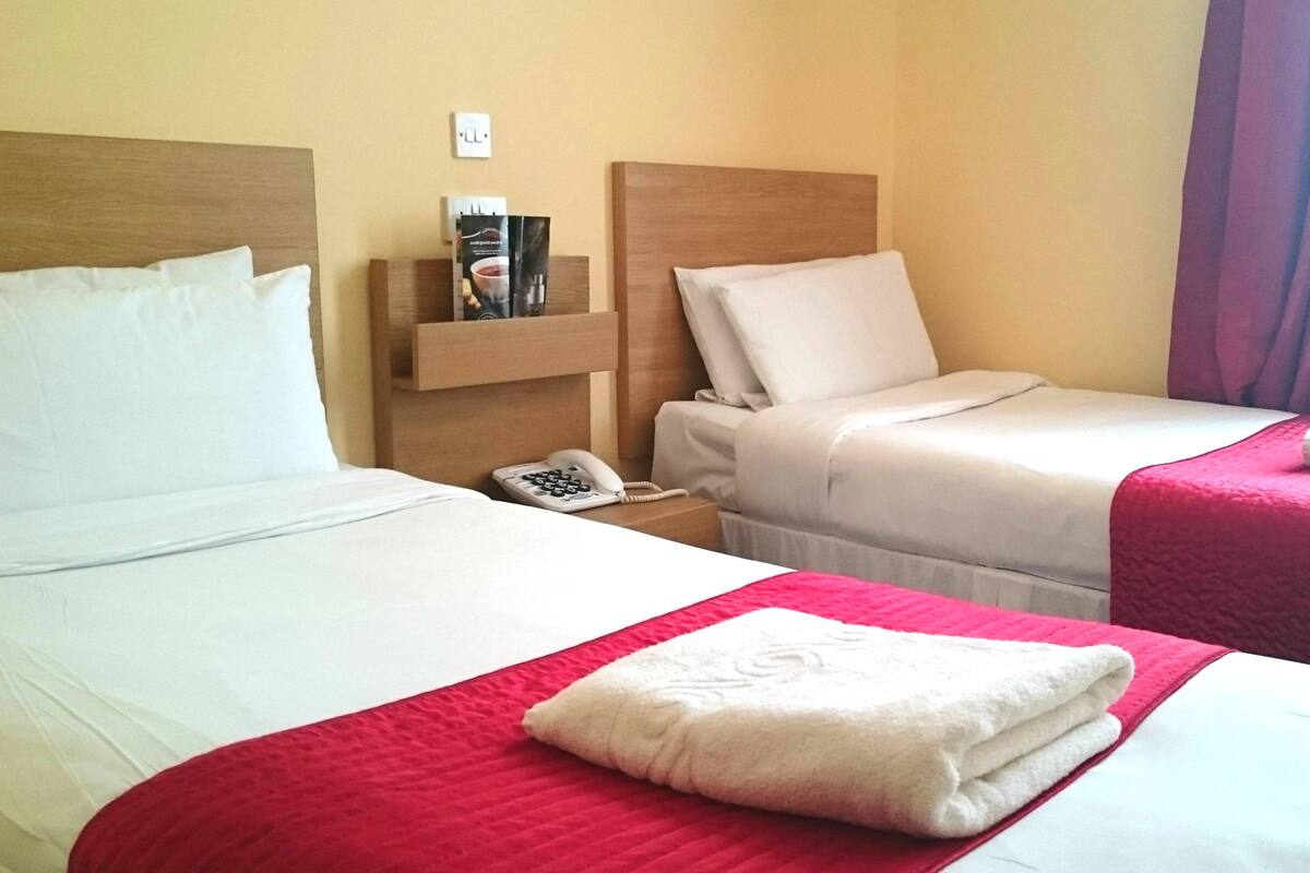 A hotel room with two beds, a nightstand with a phone, and a shelf with toiletries.