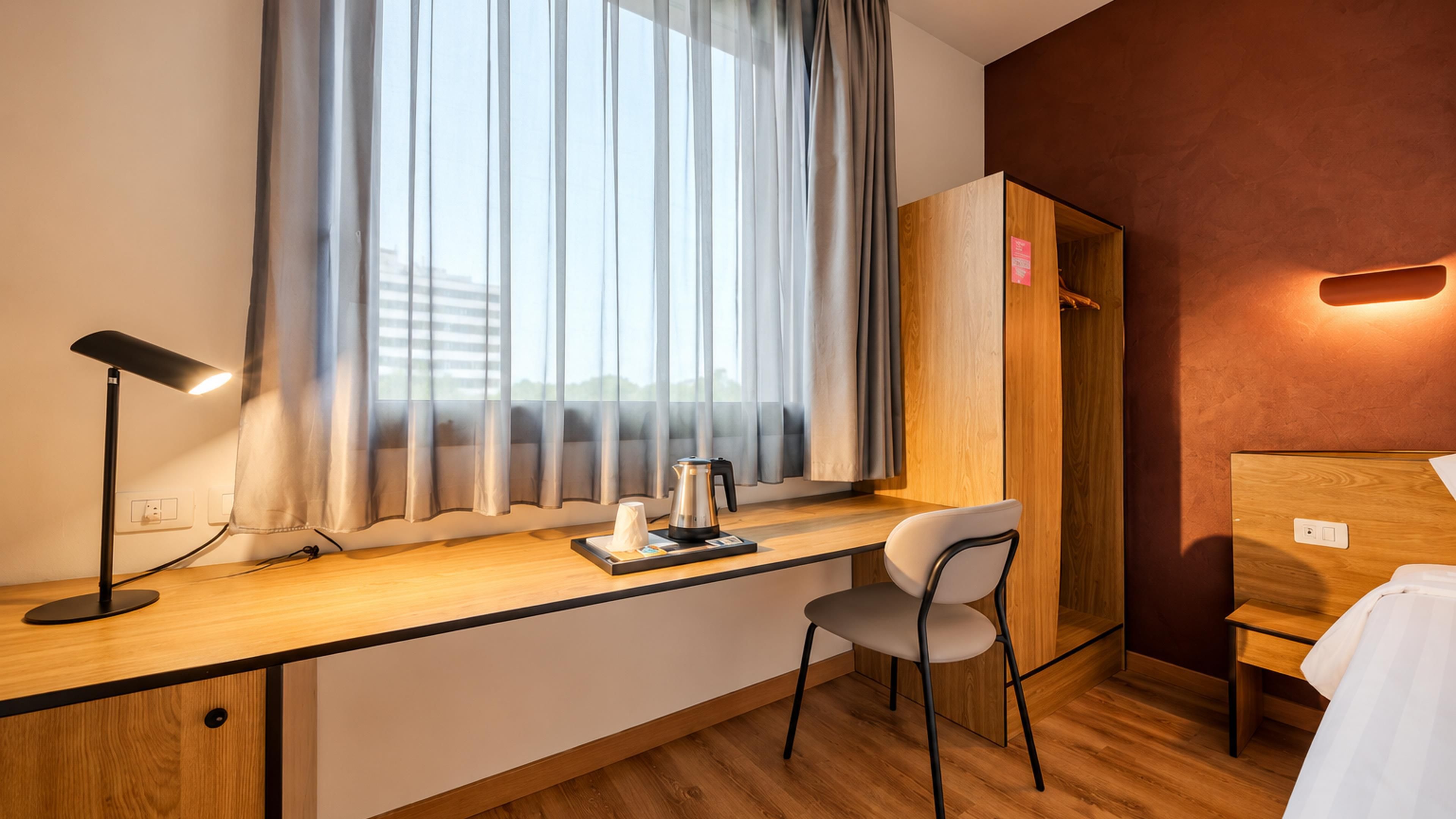 Una habitación de hotel con un escritorio de madera, una silla, una mesita de noche con lámpara, una ventana con cortinas y un armario.