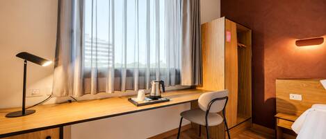 Una habitación de hotel con un escritorio de madera, una silla, una mesita de noche con lámpara, una ventana con cortinas y un armario.