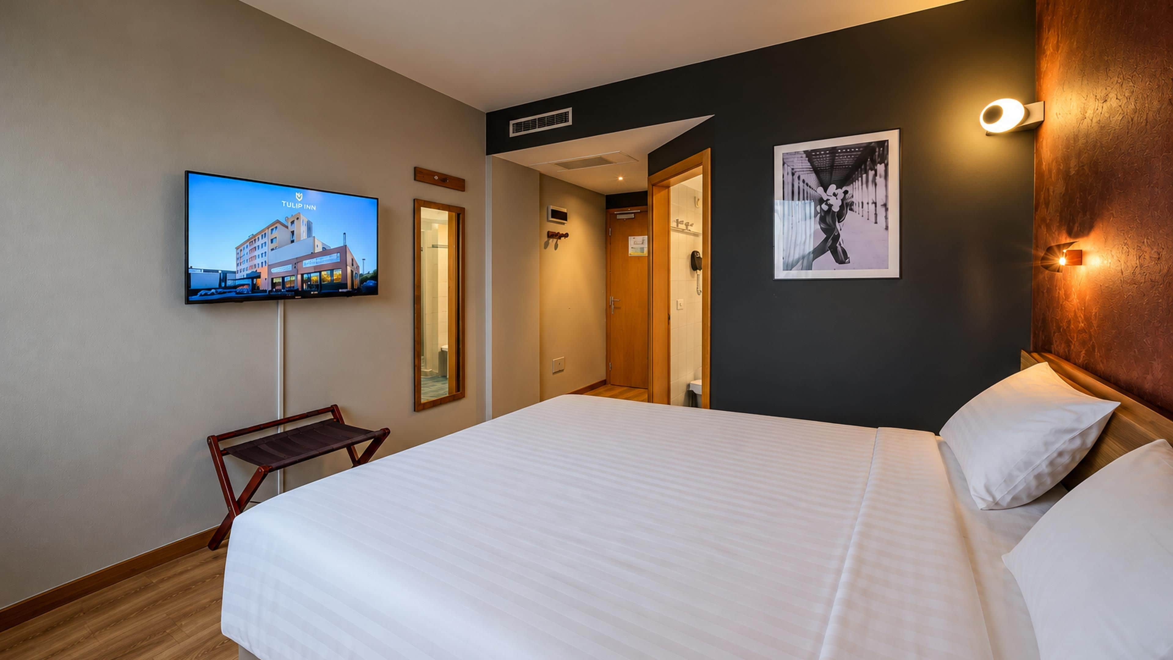 A hotel room with a bed, a TV mounted on the wall, a wooden bench, and a framed picture.