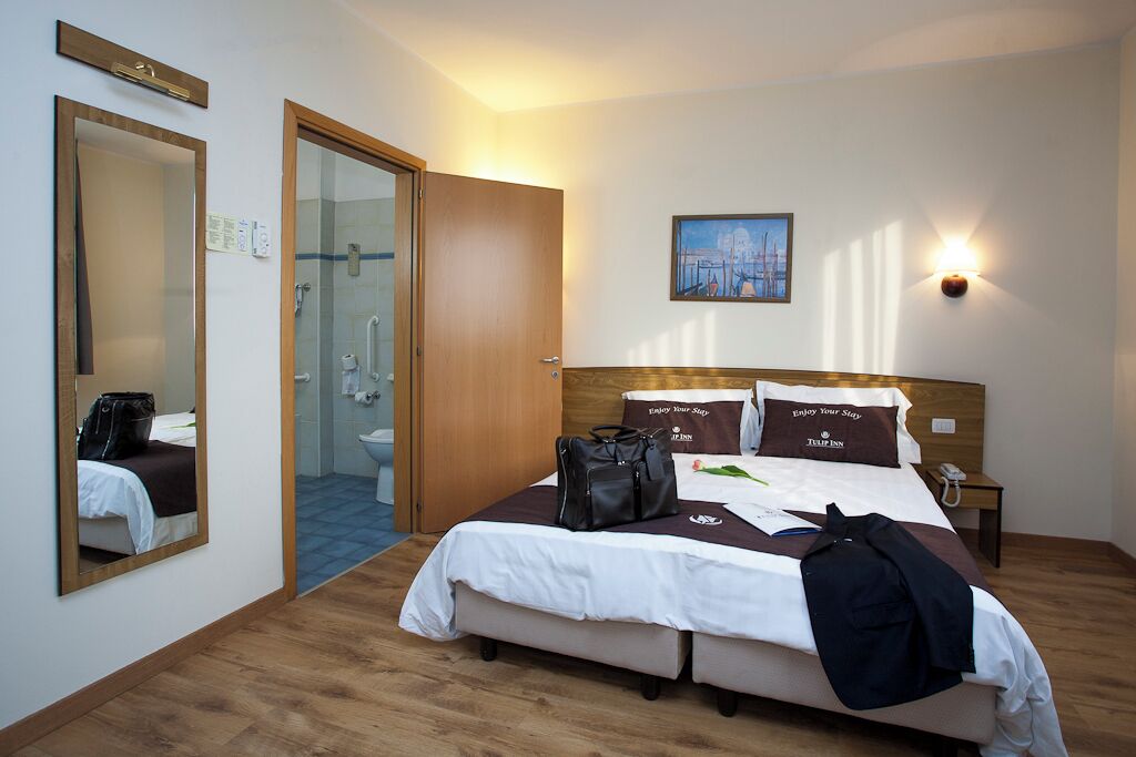 A hotel room with a bed, a bathroom, a mirror, and a painting on the wall.