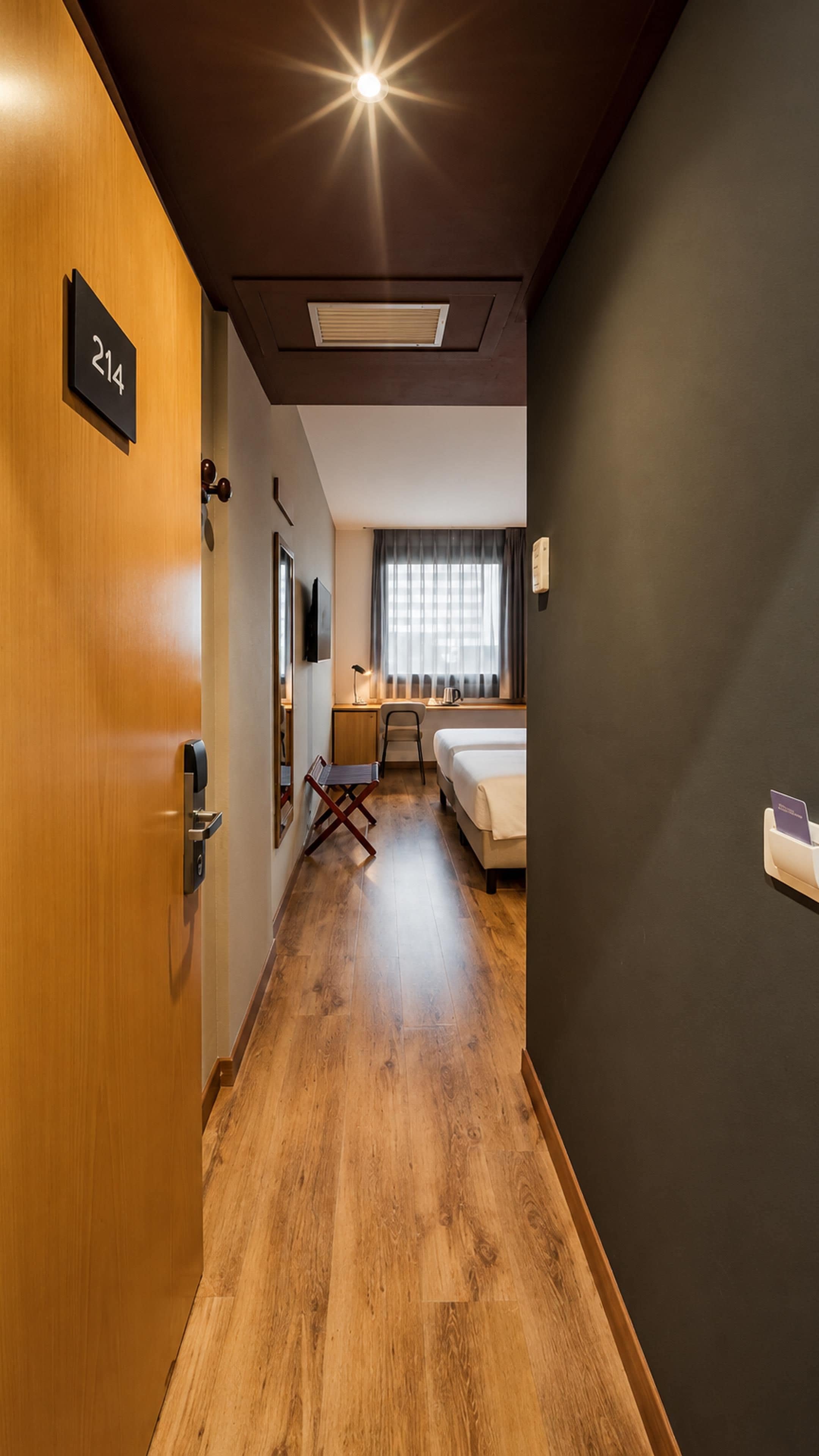 A hotel room with a wooden floor, a bed, a desk with a chair, a window with curtains, and a door with a number sign.