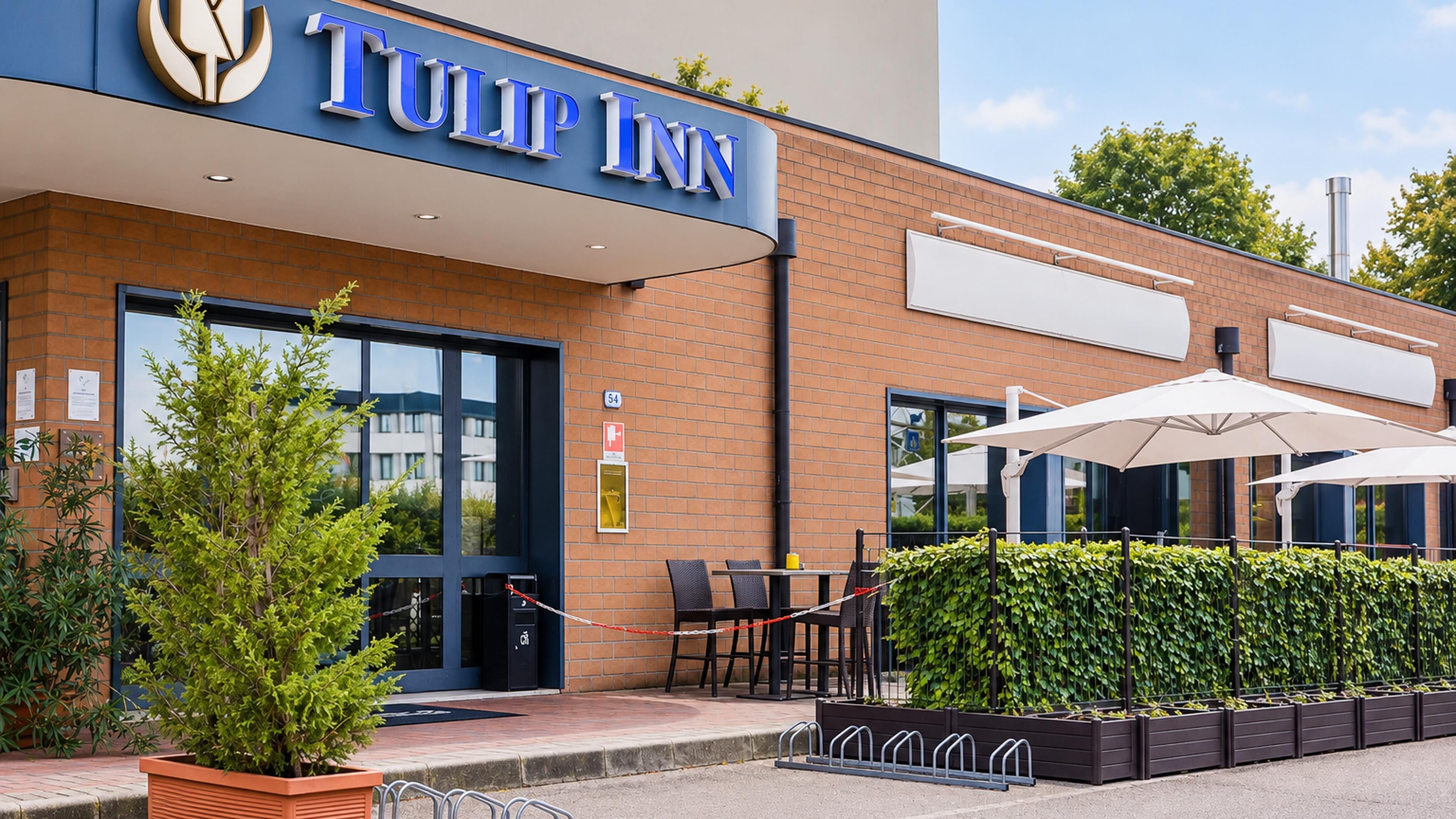 Ein Hotel Eingang mit einem Schild mit der Aufschrift 'TULIP INN', Sitzgelegenheiten im Freien inklusive Sonnenschirmen und einer gepflegten Hecke.