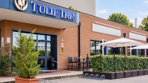 Entrada de un hotel con un letrero que dice 'TULIP INN', mobiliario exterior con sombrillas y un seto bien cuidado.