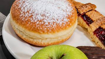 Ein Frühstücksteller mit Rührei, Speck, einem Krapfen, einem Stück Kuchen, einem Glas Orangensaft und einem Apfel.
