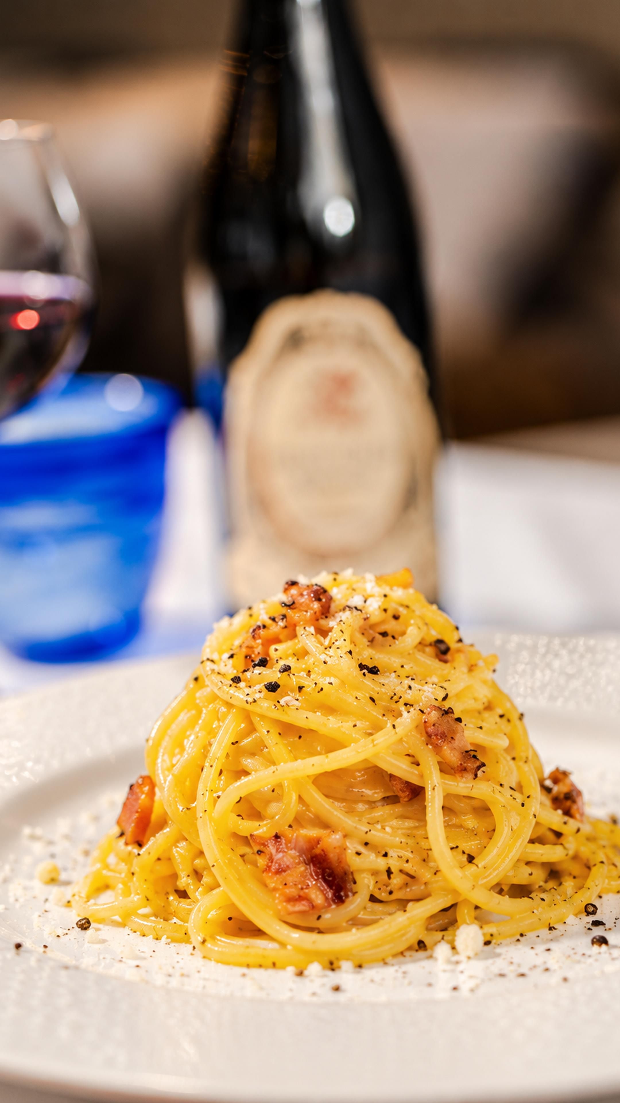 A plate of spaghetti with a creamy sauce, garnished with black pepper and cheese, with a bottle and glass in the background.
