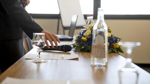 Een vergadertafel met een laptop, microfoon, glas en een bloemstuk.