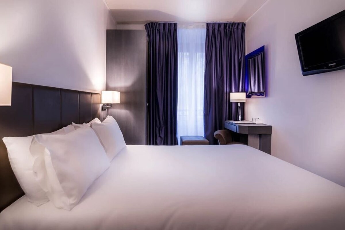 A hotel room with a large bed, bedside lamps, a desk with a chair, a flat-screen TV, and a window with curtains.