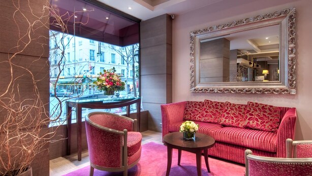 A room with a red sofa, a round coffee table, and a chair. There is a large mirror on the wall and a window with a view of buildings. - Masséna Hôtel (Paris)