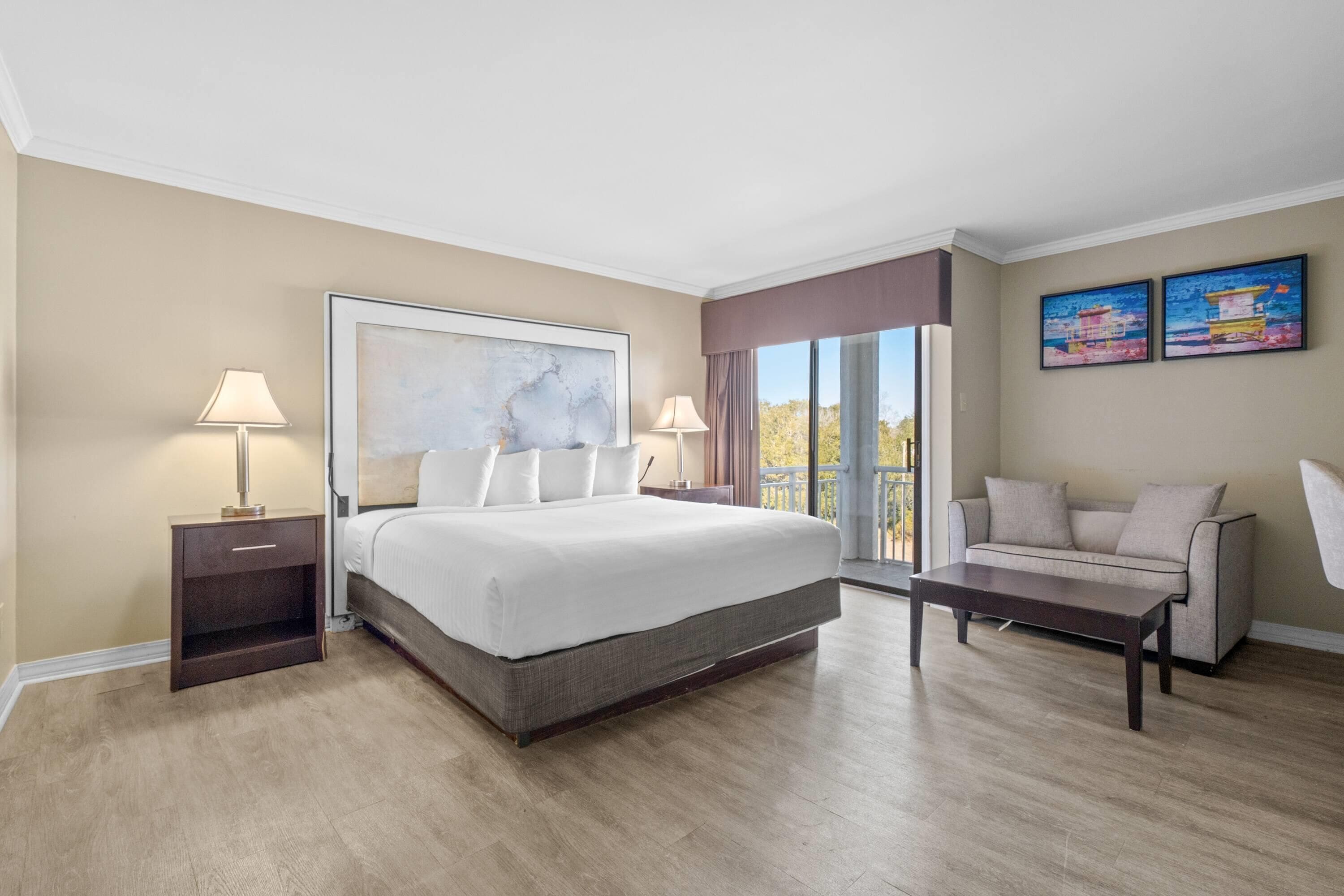 A hotel room with a large bed, two bedside tables with lamps, a sofa, and a balcony with a view.