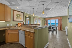 A kitchen with wooden cabinets, a dishwasher, a dining area with a table and chairs, and a view of the ocean through large windows. - The Sandbar Hotel, Trademark Collection by Wyndham (Myrtle Beach)