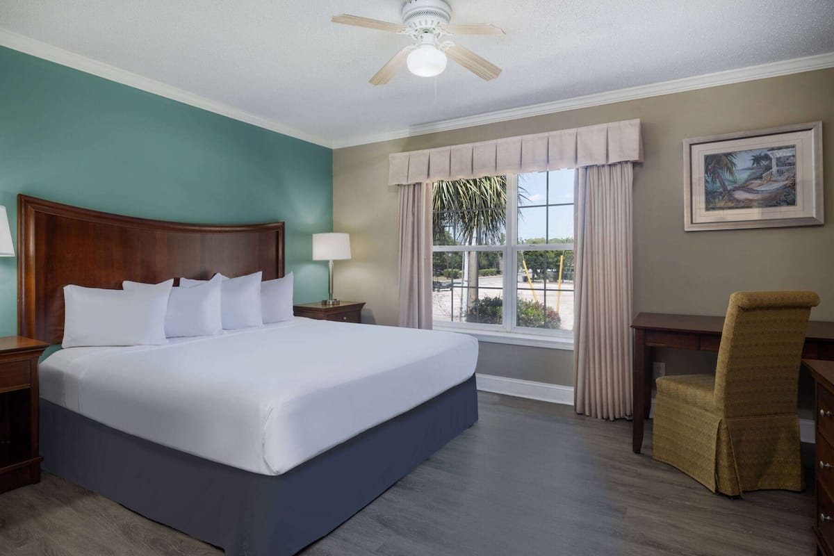 Una habitación de hotel con una cama grande, un cabecero de madera, un escritorio con una silla, una ventana con cortinas y un ventilador de techo.