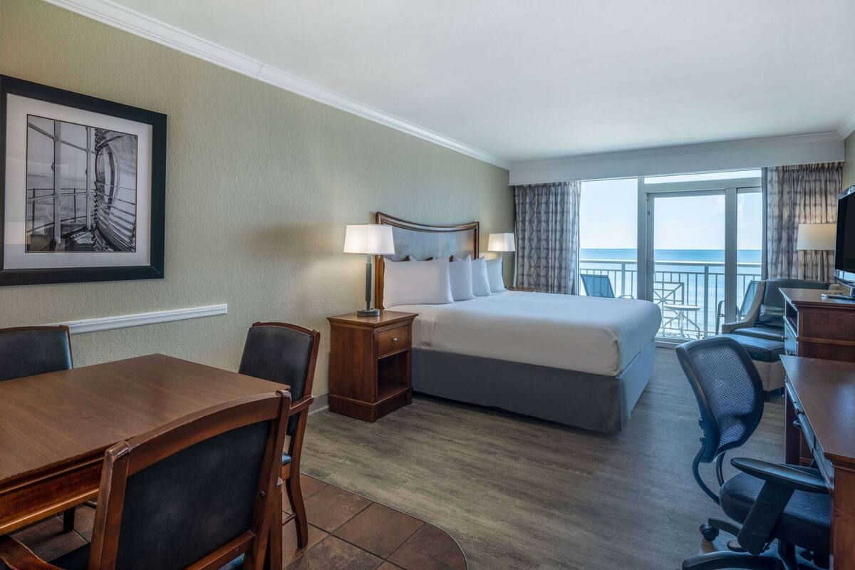 Habitación de hotel con una cama, dos mesitas de noche con lámparas, un escritorio con una silla, un cuadro enmarcado en la pared y vistas al mar a través de amplios ventanales.