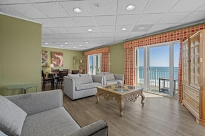 A living room with a sofa, a glass-top coffee table, and a view of the ocean through large windows. - The Sandbar Hotel, Trademark Collection by Wyndham (Myrtle Beach)