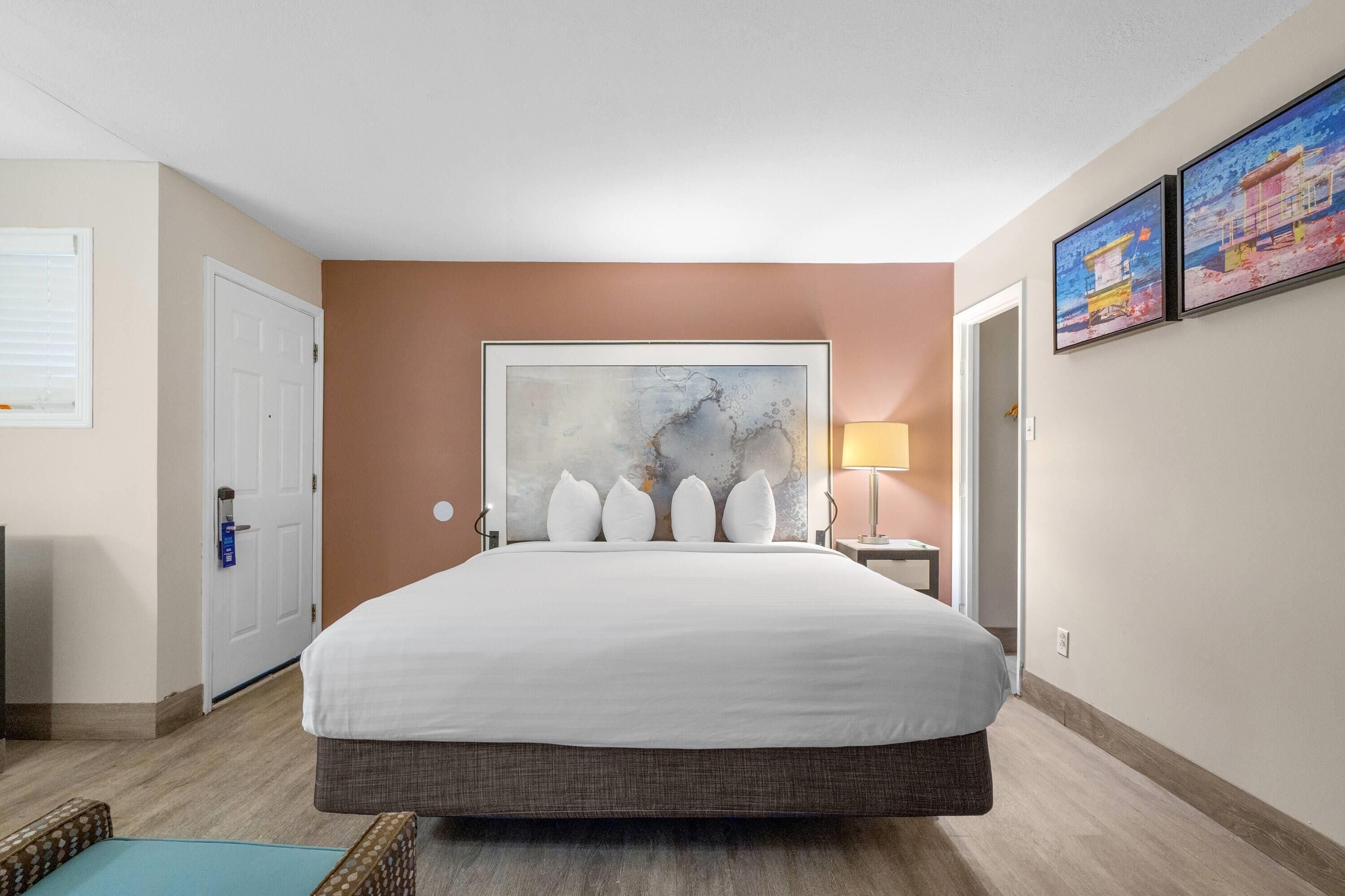 Ein Hotelzimmer mit einem großen Bett, einer Tür, einem Wandbild und einem Nachttisch mit Lampe.