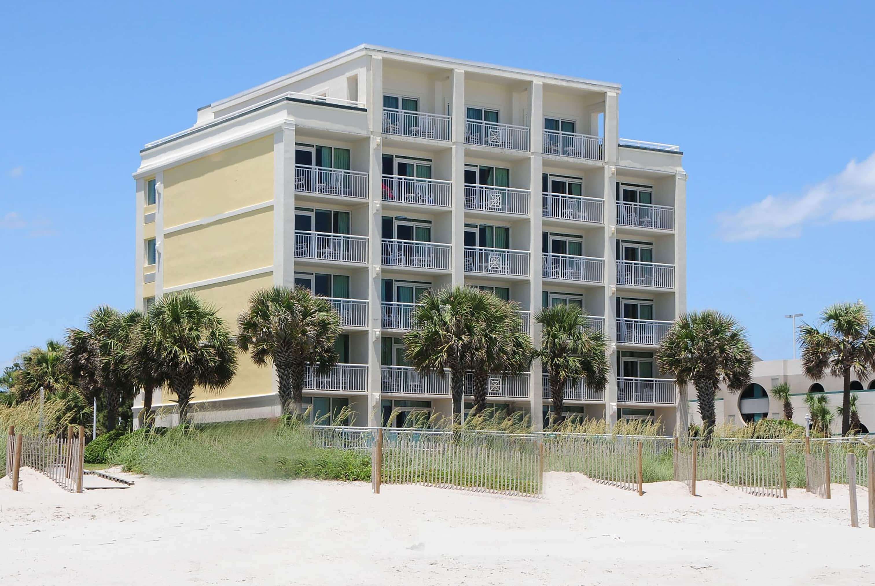 Een hotel met meerdere verdiepingen en balkons, dichtbij een zandstrand met palmbomen.