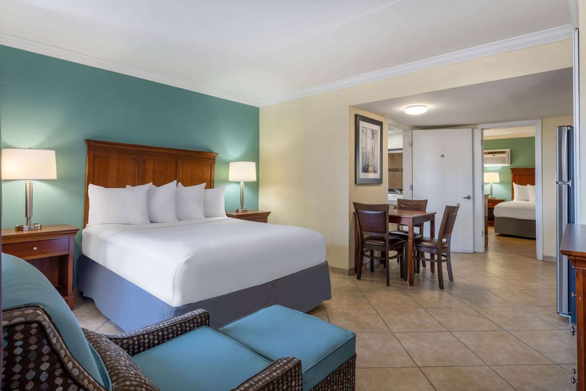 Habitación de hotel con una cama grande, dos mesitas de noche con lámparas, una pequeña mesa de comedor con sillas y una zona de kitchenette.