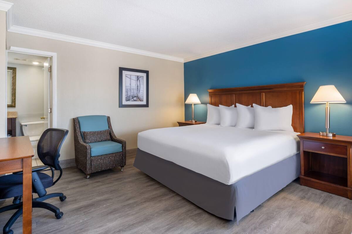 Habitación de hotel con cama, dos lámparas de noche, una silla, un escritorio y un cuadro en la pared.