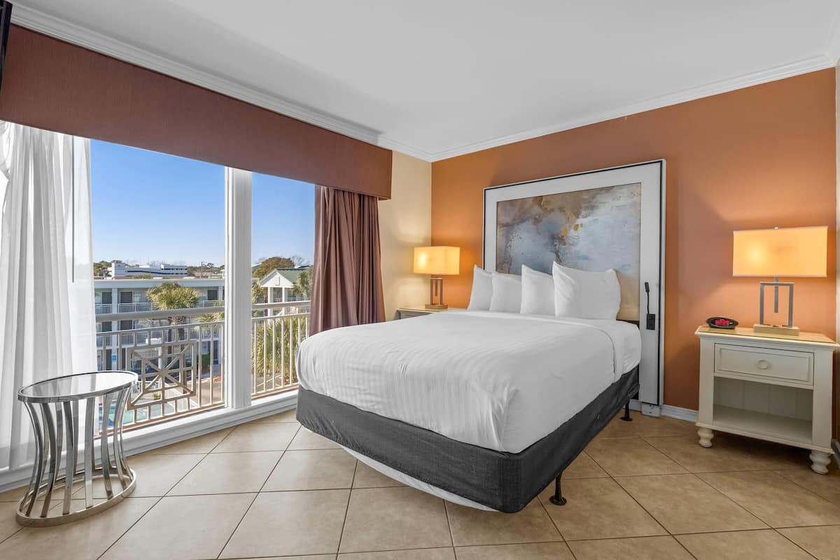 Una habitación de hotel con una cama grande, dos mesitas de noche, vistas a la ciudad y un balcón con piscina.