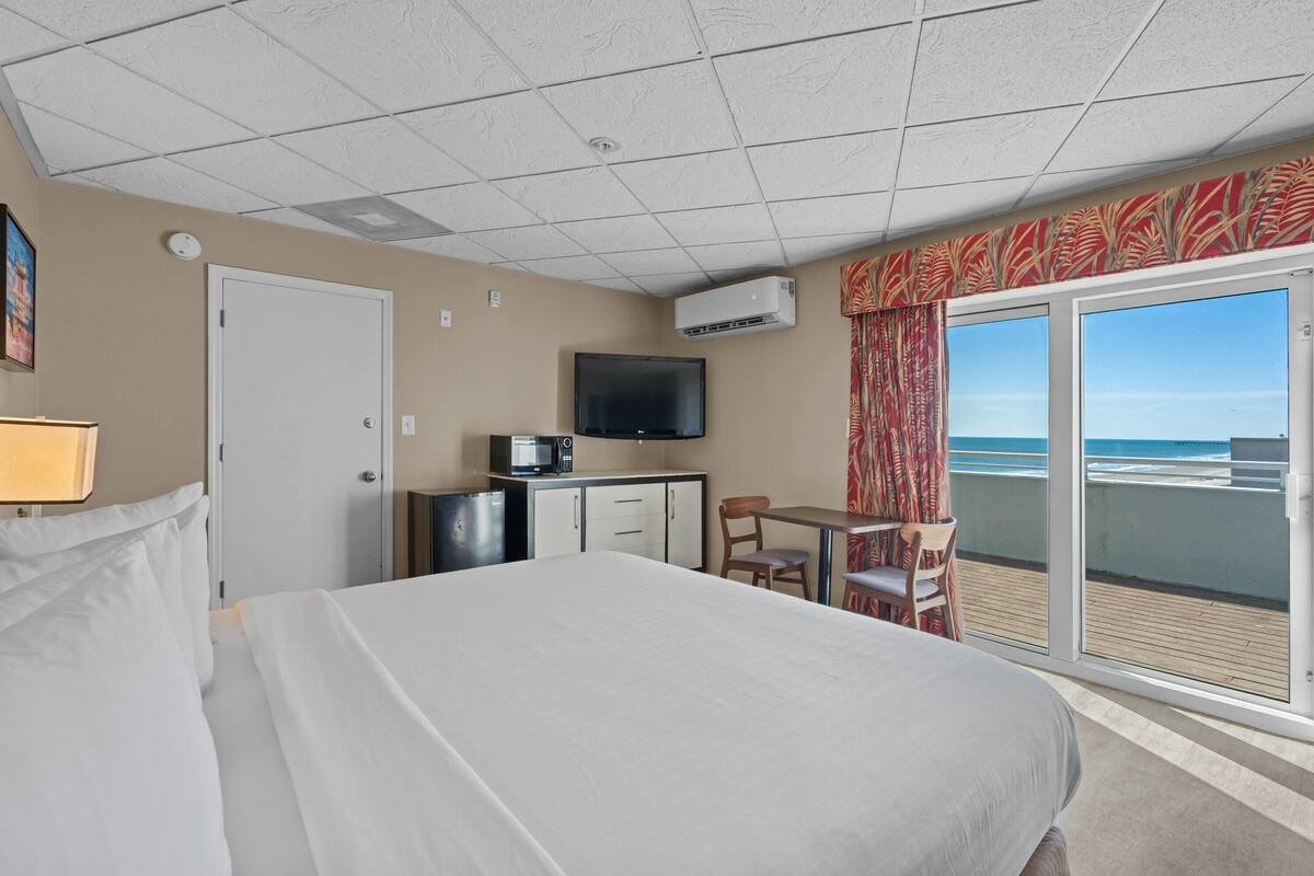 Habitación de hotel con cama, escritorio con televisor, cafetera, refrigerador pequeño y puerta corrediza de vidrio al exterior.