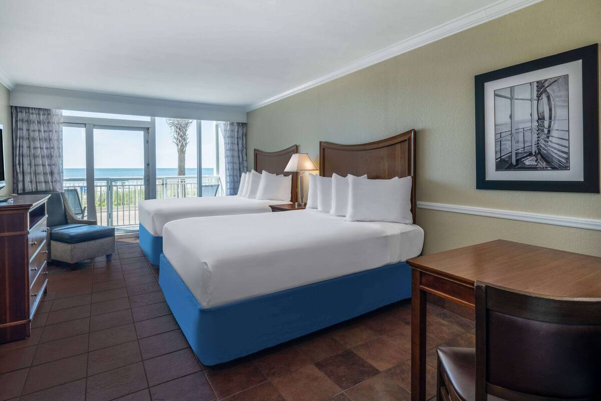 Habitación de hotel con dos camas, un escritorio, una silla, un televisor y un balcón con vista al mar.