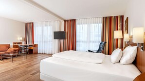 A hotel room with a large bed, a desk with a chair, a TV, and a sofa. - NH München Ost Conference Center (Aschheim)