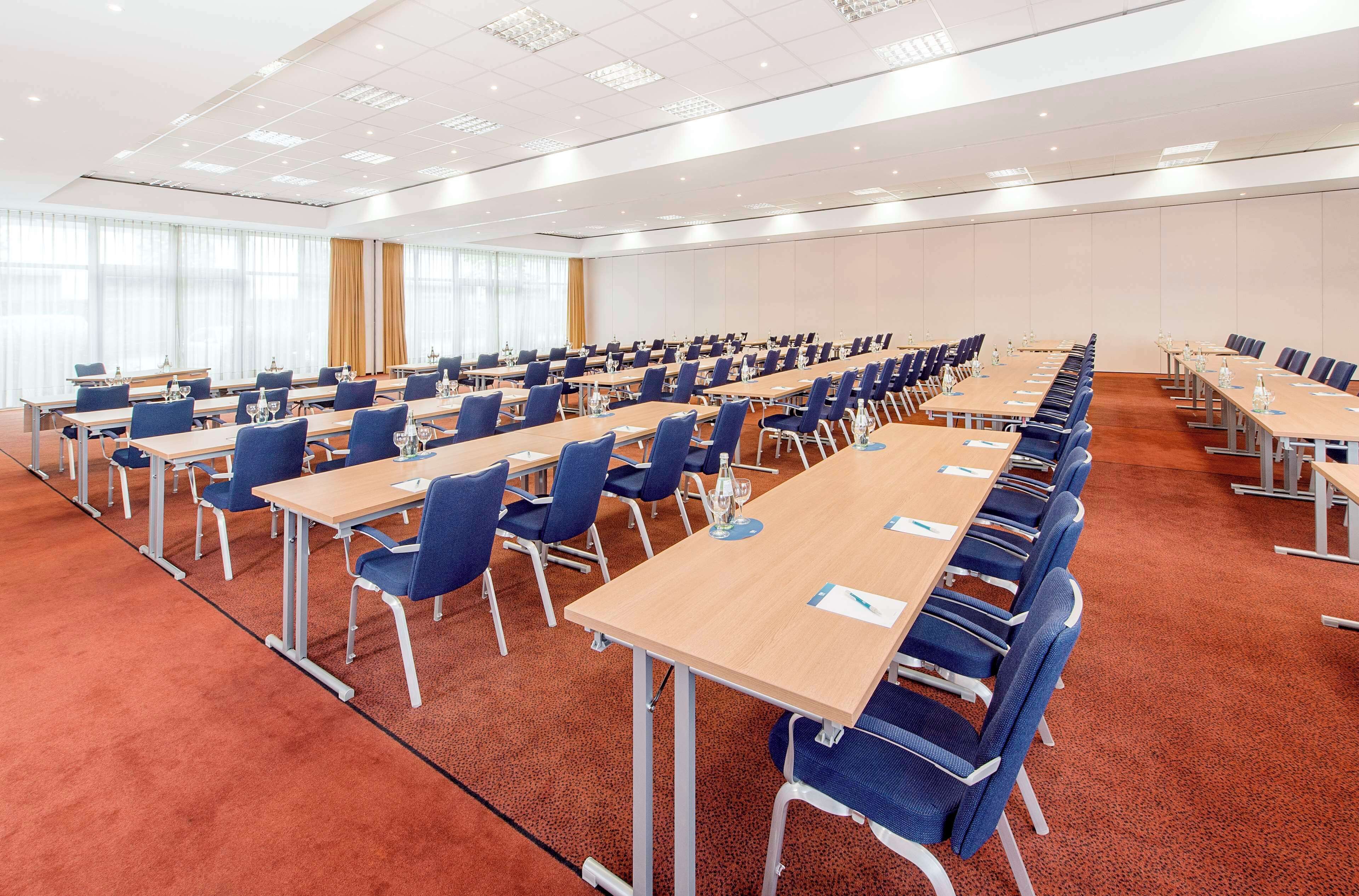 A large conference room with rows of tables and chairs, carpeted floor, and large windows with curtains.